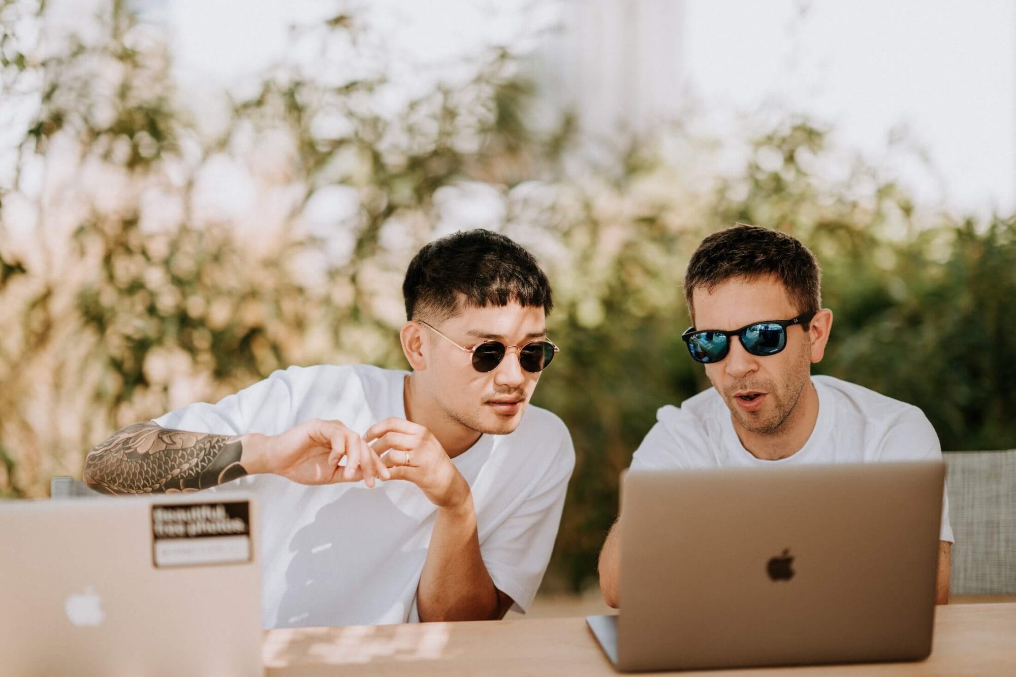 Deux hommes en t-shirts blancs et lunettes de soleil travaillant sur un ordinateur portable Apple en extérieur.