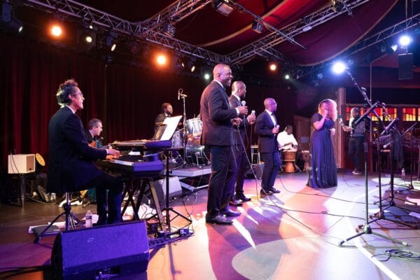 Un groupe de chanteurs et musiciens en pleine performance sur une scène illuminée. Un pianiste et un guitariste accompagnent les chanteurs, tandis qu’un batteur et un percussionniste jouent en arrière-plan.