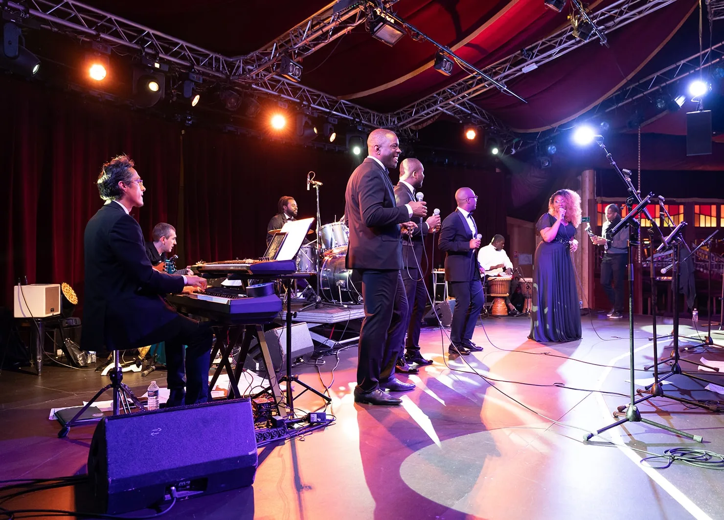 Un groupe de chanteurs et musiciens en pleine performance sur une scène illuminée. Un pianiste et un guitariste accompagnent les chanteurs, tandis qu’un batteur et un percussionniste jouent en arrière-plan.