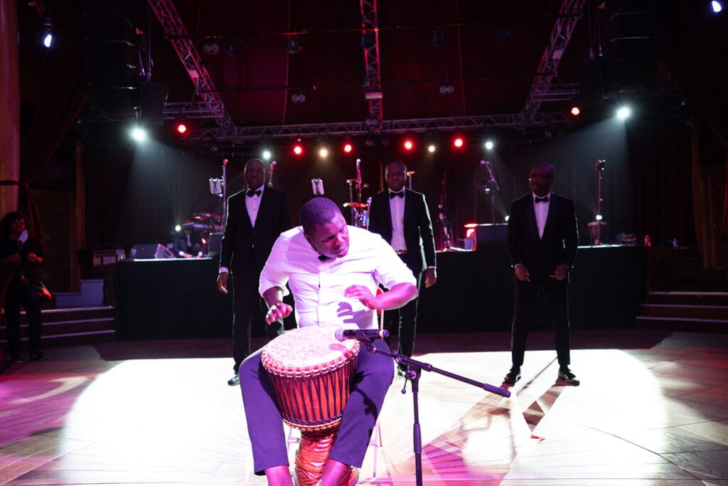 Un percussionniste joue du djembé avec intensité sur une scène éclairée de lumières rouges et blanches, tandis que quatre hommes en costume noir et nœud papillon se tiennent derrière lui.