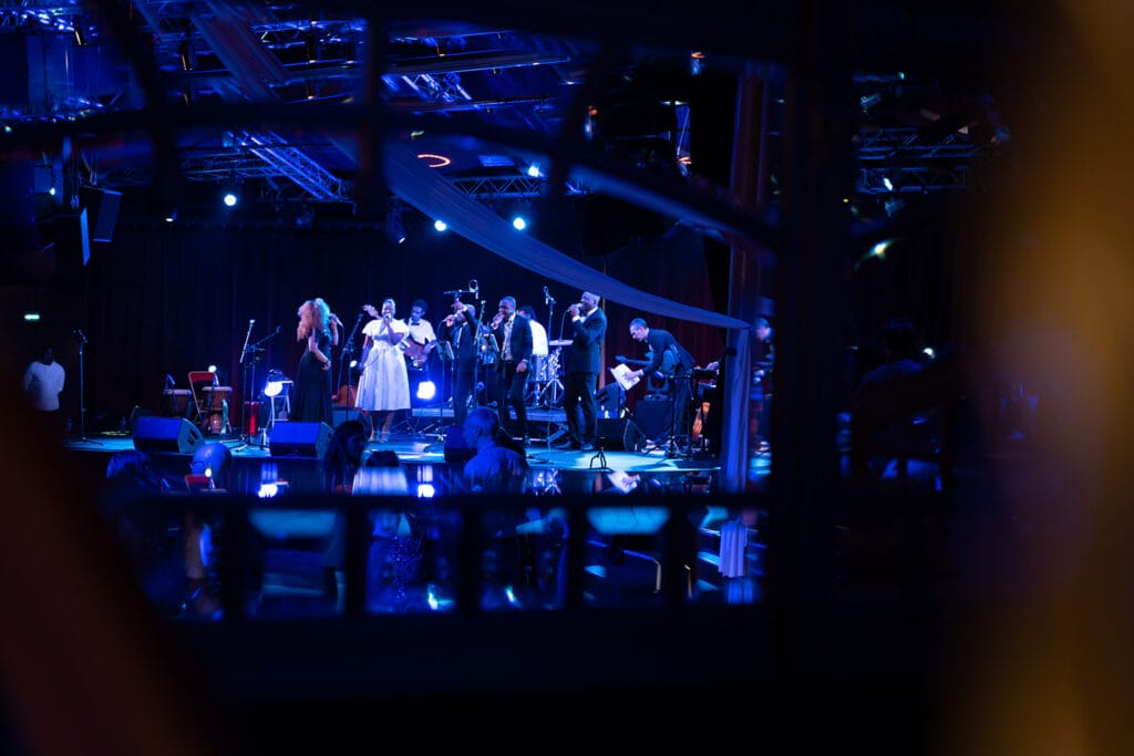 Un groupe de musiciens et chanteurs se produit sur une scène illuminée de lumières bleues, créant une ambiance immersive devant un public attentif.