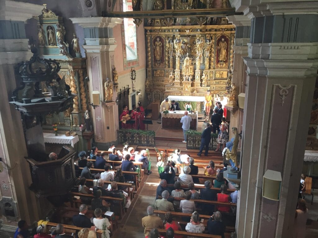 Chorale lors d'une cérémonie religieuse : un musicien en robe de chœur rose joue du clavier, entouré de participants masqués, debout et applaudissant dans une église ornée de décorations anciennes.