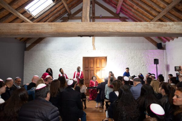 Des membres d'une chorale gospel vêtus de robes rouges et blanches chantent devant un public attentif dans une salle lumineuse avec un toit en bois.