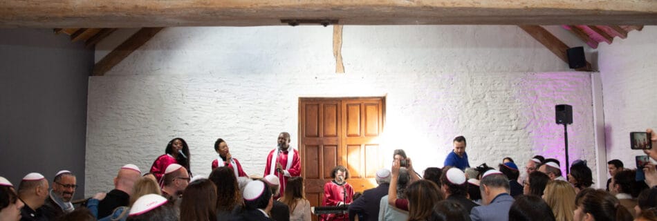 Des membres d'une chorale gospel vêtus de robes rouges et blanches chantent devant un public attentif dans une salle lumineuse avec un toit en bois.