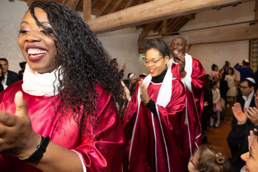 Des chanteurs gospel vêtus de robes rouges et blanches avancent en souriant et en applaudissant parmi le public lors d'un événement festif.