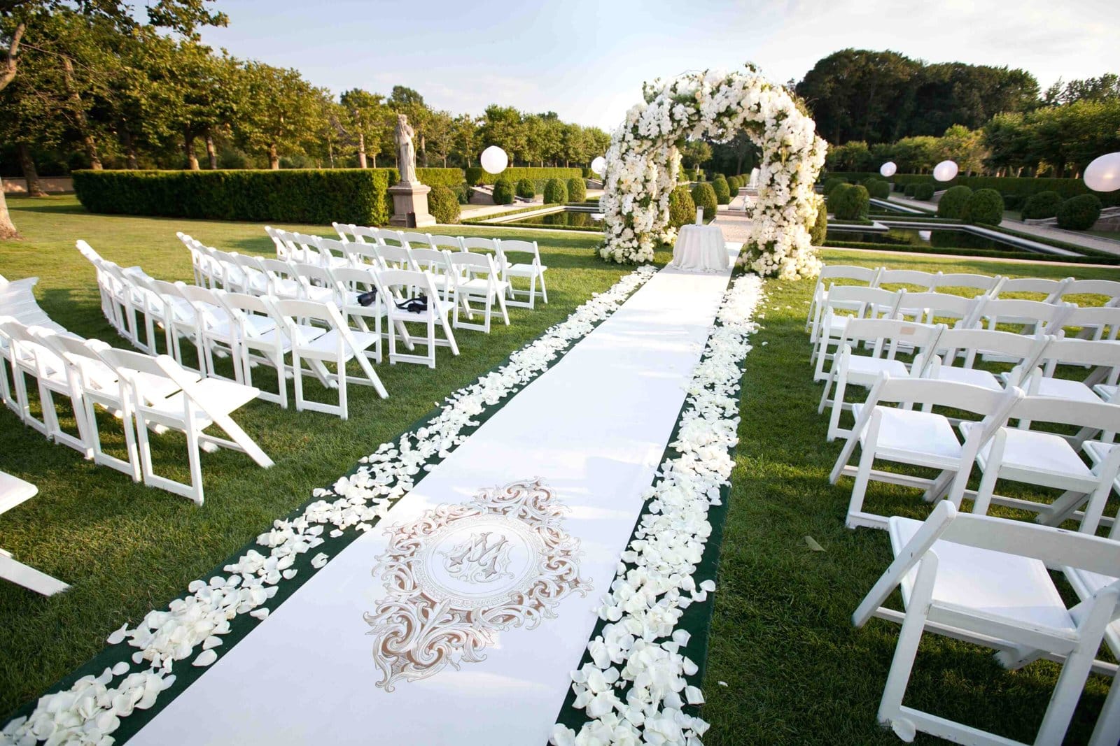 Allée de mariage décorée de pétales de roses blanches menant à une arche florale dans un jardin verdoyant.