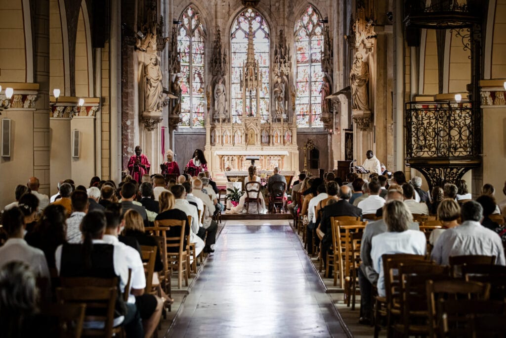 Vue arrière d’une église majestueuse remplie d’invités assistant à une cérémonie de mariage, avec les mariés assis face à l’autel et un chœur gospel en tenue rose chantant à leurs côtés.