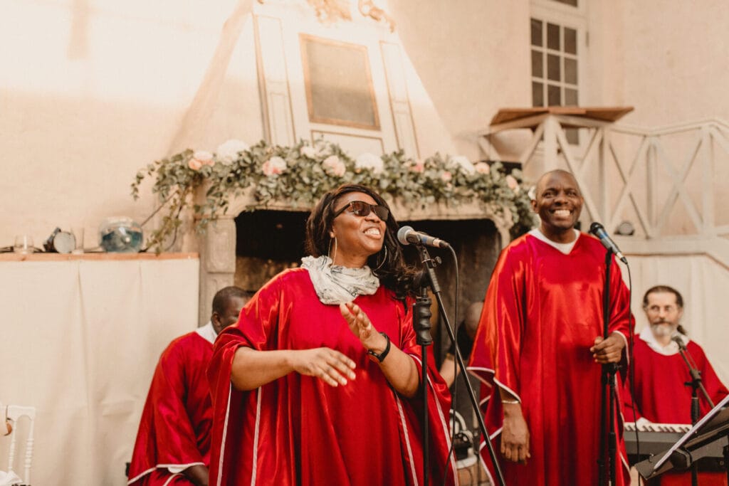 Une chanteuse gospel en robe rose satinée chante avec passion dans un micro, accompagnée de choristes, lors d'une cérémonie dans une église richement décorée.