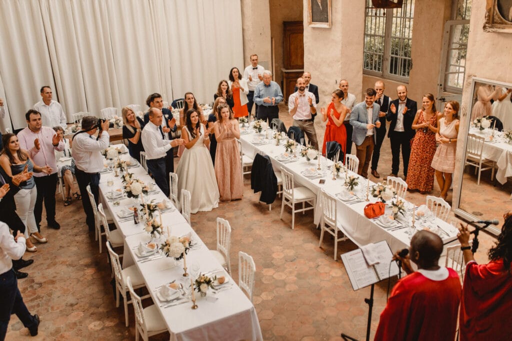 Invités réunis sous une pergola pour un cocktail de mariage élégant en extérieur, entourés de verdure et d’une bâtisse en pierre.