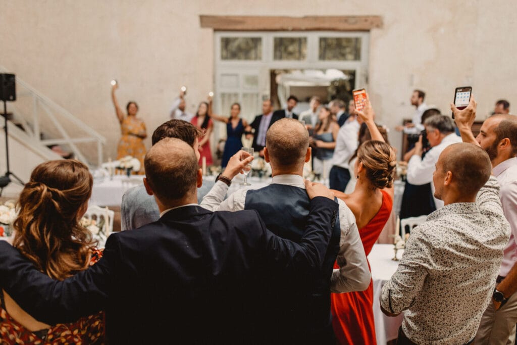 Vue de dos d’un groupe d’invités levant leurs verres et leurs téléphones, tandis qu’en face, d’autres convives chantent et lèvent les bras. L’ambiance est festive et émotive, capturant un moment de communion joyeuse.