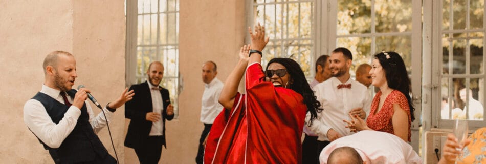 Un moment de fête intense lors d’un mariage. Un chanteur en costume chante avec énergie dans un micro, tandis qu’une femme en robe rouge applaudit en dansant. En arrière-plan, d’autres invités s’amusent et profitent de l’ambiance conviviale sous la lumière naturelle d’une salle aux grandes fenêtres.