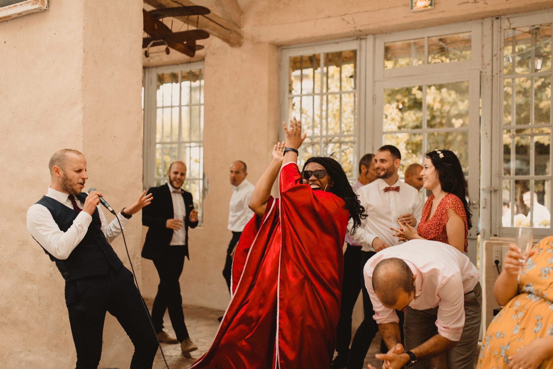 Un moment de fête intense lors d’un mariage. Un chanteur en costume chante avec énergie dans un micro, tandis qu’une femme en robe rouge applaudit en dansant. En arrière-plan, d’autres invités s’amusent et profitent de l’ambiance conviviale sous la lumière naturelle d’une salle aux grandes fenêtres.