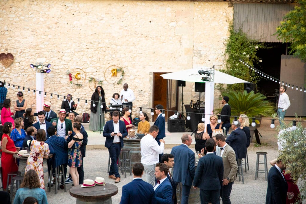 Une réception de mariage en extérieur, où des invités en tenue élégante discutent et trinquent autour de tonneaux aménagés en tables. Un groupe musical se produit sur une petite scène contre un mur en pierre décoré de guirlandes et de lettres dorées. L’ambiance est festive et romantique.