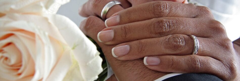 Mains d’un couple de mariés portant leurs alliances, entrelacées avec tendresse, accompagnées d’un bouquet de roses blanches.