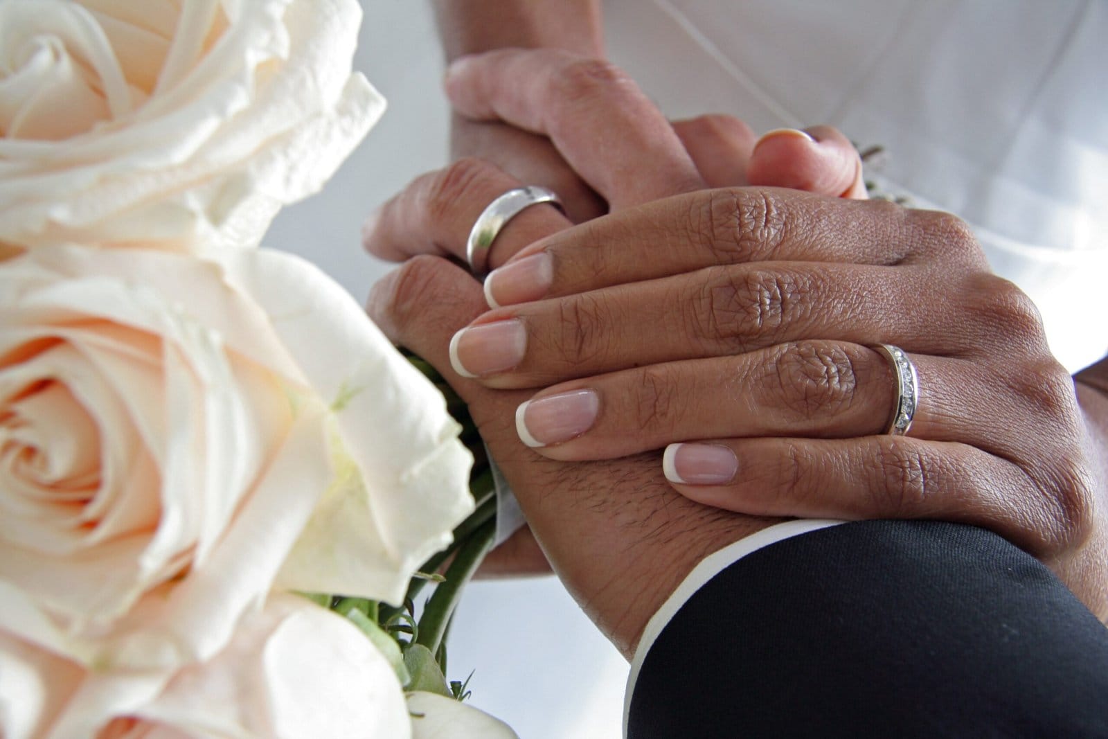 Mains d’un couple de mariés portant leurs alliances, entrelacées avec tendresse, accompagnées d’un bouquet de roses blanches.