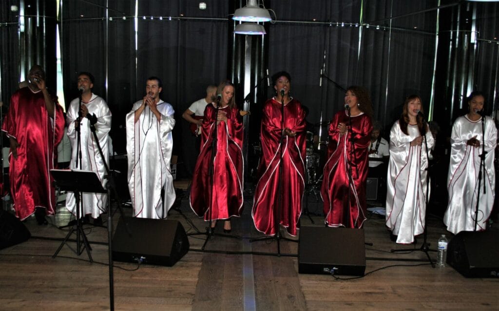 Une chorale gospel en robe de cérémonie rouge et blanche chante sur scène, entourée de musiciens. L’expression intense des chanteurs et la profondeur de leur interprétation captivent le public, offrant une expérience musicale authentique et émouvante.