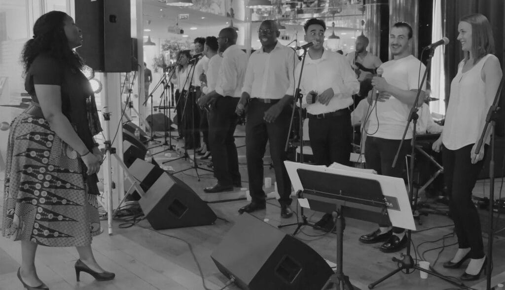 Un groupe de musiciens et chanteurs se produit sur une scène illuminée de lumières bleues, créant une ambiance immersive devant un public attentif.