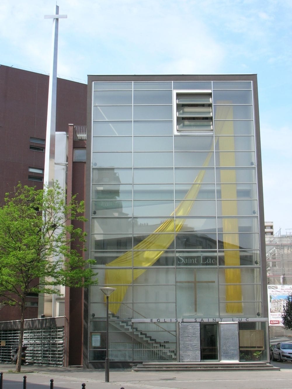 L’image montre une église moderne nommée "Église Saint Luc". Son architecture est contemporaine, avec une grande façade en verre qui laisse entrevoir l’intérieur. Une croix métallique est érigée à gauche du bâtiment, et un tissu jaune orne l'intérieur, créant un effet visuel marquant. L’entrée se situe en bas, avec des portes vitrées et une inscription indiquant le nom de l’église.