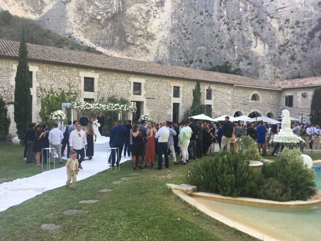 Vue d’une cérémonie de mariage en plein air sous une pergola en bois, avec des invités assis sur des chaises blanches et un chœur gospel vêtu de rouge. L’ambiance est chaleureuse, décorée de lanternes et de pétales de fleurs au sol.