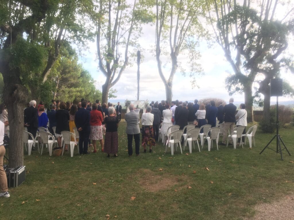 Vue d’une cérémonie de mariage en plein air sous une pergola en bois, avec des invités assis sur des chaises blanches et un chœur gospel vêtu de rouge. L’ambiance est chaleureuse, décorée de lanternes et de pétales de fleurs au sol.