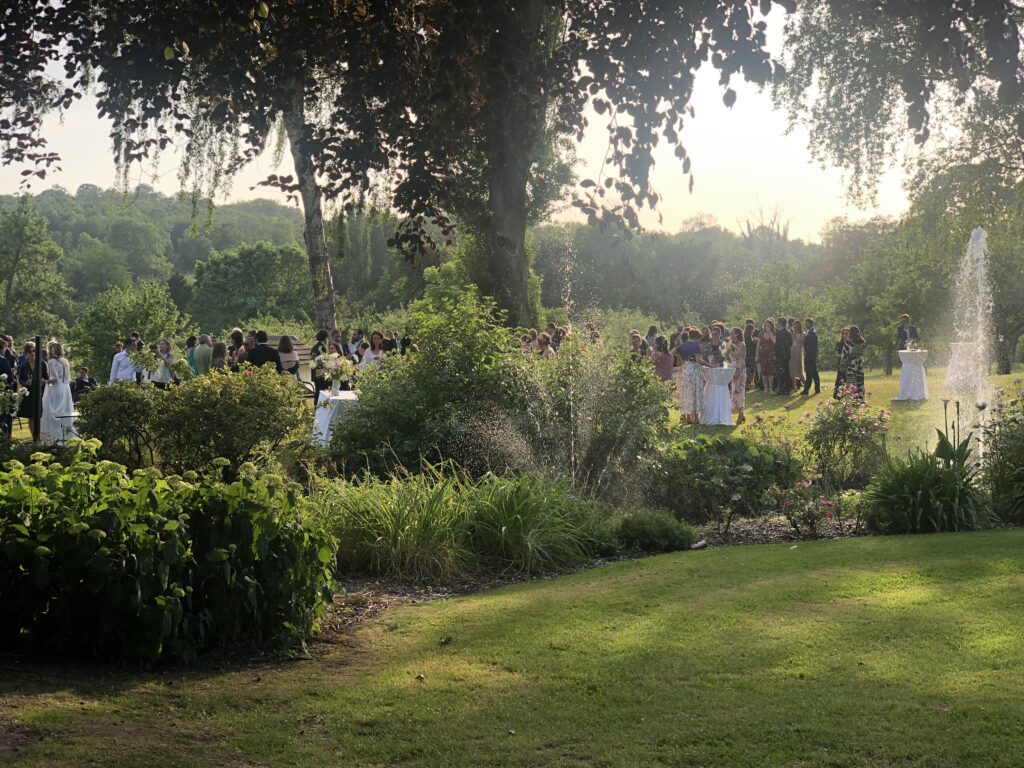 Une réception de mariage dans un lieu au charme ancien, avec des invités élégamment vêtus applaudissant la mariée en robe blanche, tandis qu'un groupe en tenue rouge anime l'événement.