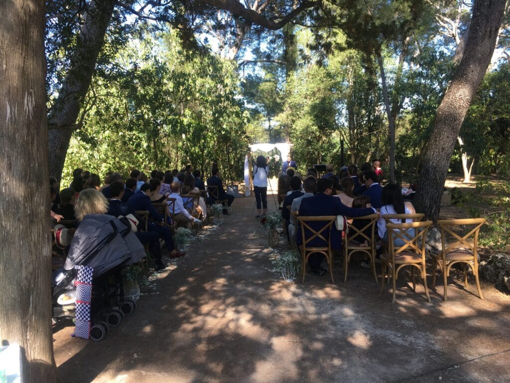 Une cérémonie de mariage en extérieur, sous l’ombre des arbres, avec des invités assis sur des chaises en bois, face aux mariés et à l’officiant. L’atmosphère est intime et chaleureuse, en parfaite harmonie avec la nature environnante.