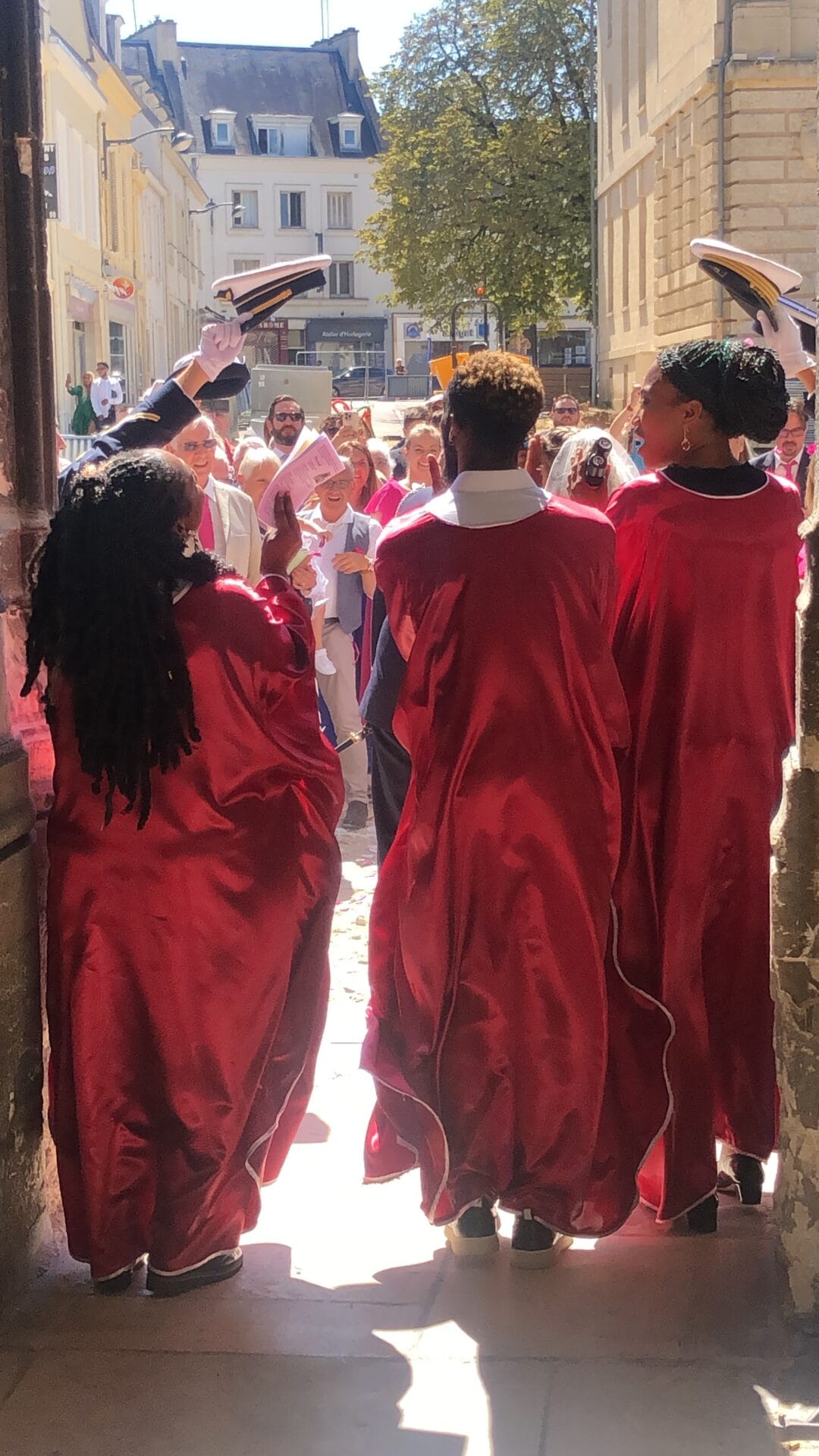 Trois choristes en Tage rouge chantent à la sortie d’une église, accueillant les mariés avec une haie d’honneur formée par des officiers levant leurs casquettes.