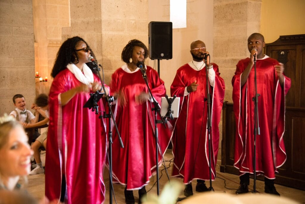 Quatre chanteurs gospel en tenue rouge et blanche interprètent une chanson lors d’une cérémonie dans une église en pierre. Leur performance passionnée crée une ambiance chaleureuse et émotive, sous le regard émerveillé des invités.