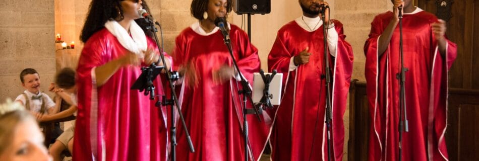 Quatre chanteurs gospel en tenue rouge et blanche interprètent une chanson lors d’une cérémonie dans une église en pierre. Leur performance passionnée crée une ambiance chaleureuse et émotive, sous le regard émerveillé des invités.