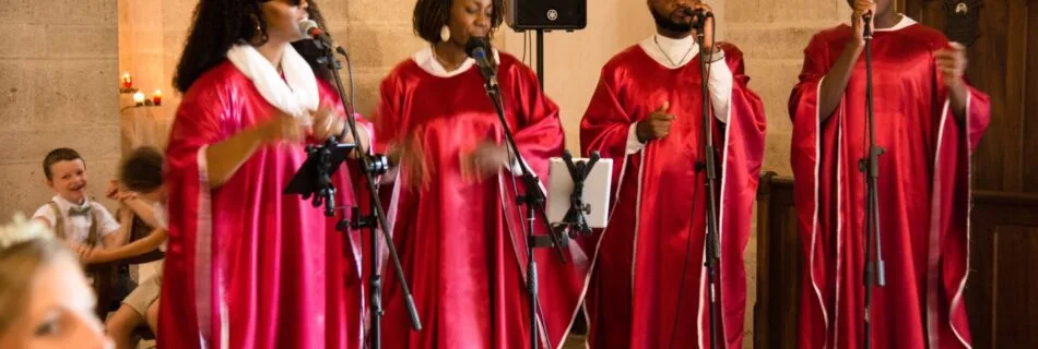 Quatre chanteurs gospel en tenue rouge et blanche interprètent une chanson lors d’une cérémonie dans une église en pierre. Leur performance passionnée crée une ambiance chaleureuse et émotive, sous le regard émerveillé des invités.
