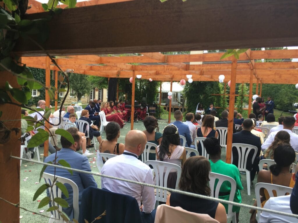 Vue d’une cérémonie de mariage en plein air sous une pergola en bois, avec des invités assis sur des chaises blanches et un chœur gospel vêtu de rouge. L’ambiance est chaleureuse, décorée de lanternes et de pétales de fleurs au sol.