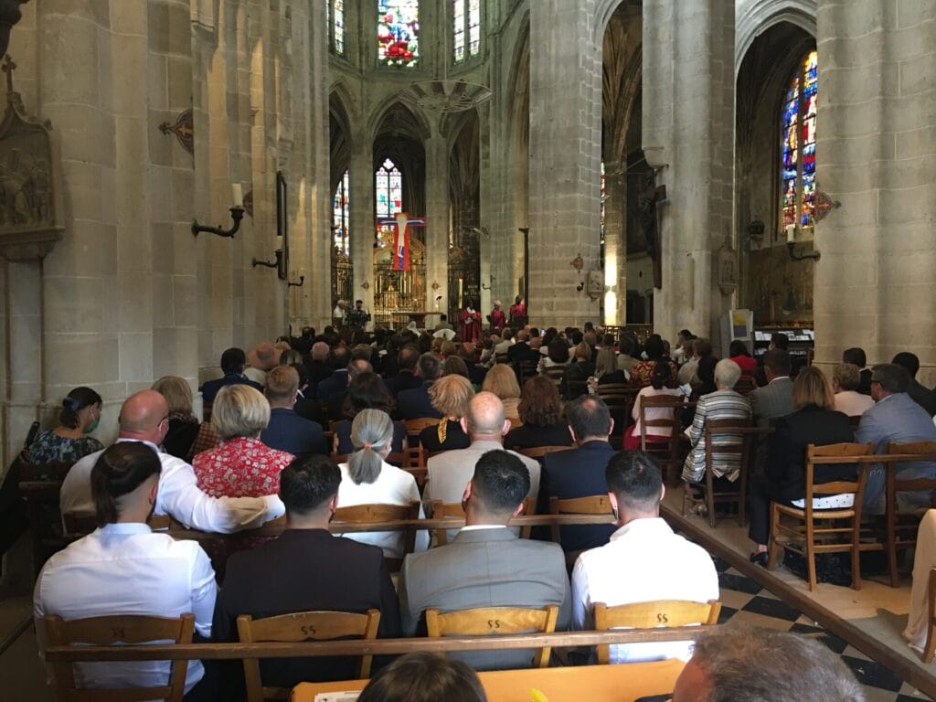 Chorale lors d'une cérémonie religieuse : un musicien en robe de chœur rose joue du clavier, entouré de participants masqués, debout et applaudissant dans une église ornée de décorations anciennes.