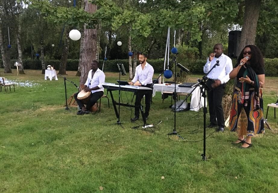 Groupe de musiciens jouant en plein air lors d'un événement, avec une chanteuse, un percussionniste, un pianiste et un chanteur.