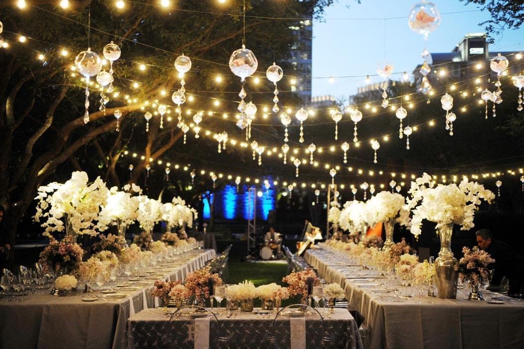 Photo d'une réception de mariage en plein air la nuit. Des guirlandes lumineuses sont suspendues entre les arbres, créant un effet de ciel étoilé. Plusieurs longues tables sont disposées avec des nappes blanches, des verres en cristal, et des centres de table composés de grandes fleurs blanches et roses. Des chaises élégantes sont placées autour des tables, et des invités sont assis, profitant de l'ambiance festive.