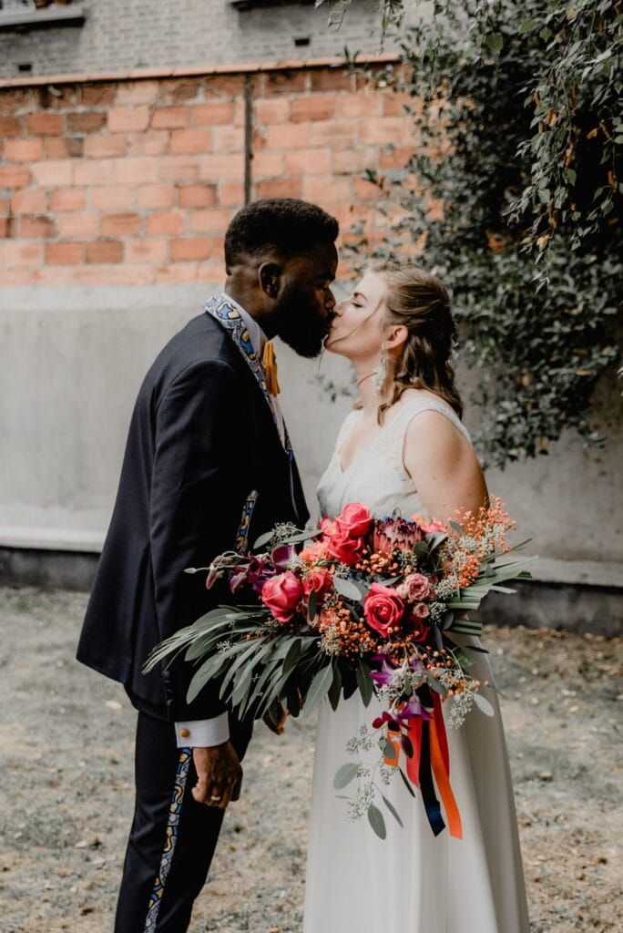 Un couple de mariés échange un tendre baiser en extérieur, la mariée tenant un grand bouquet coloré.