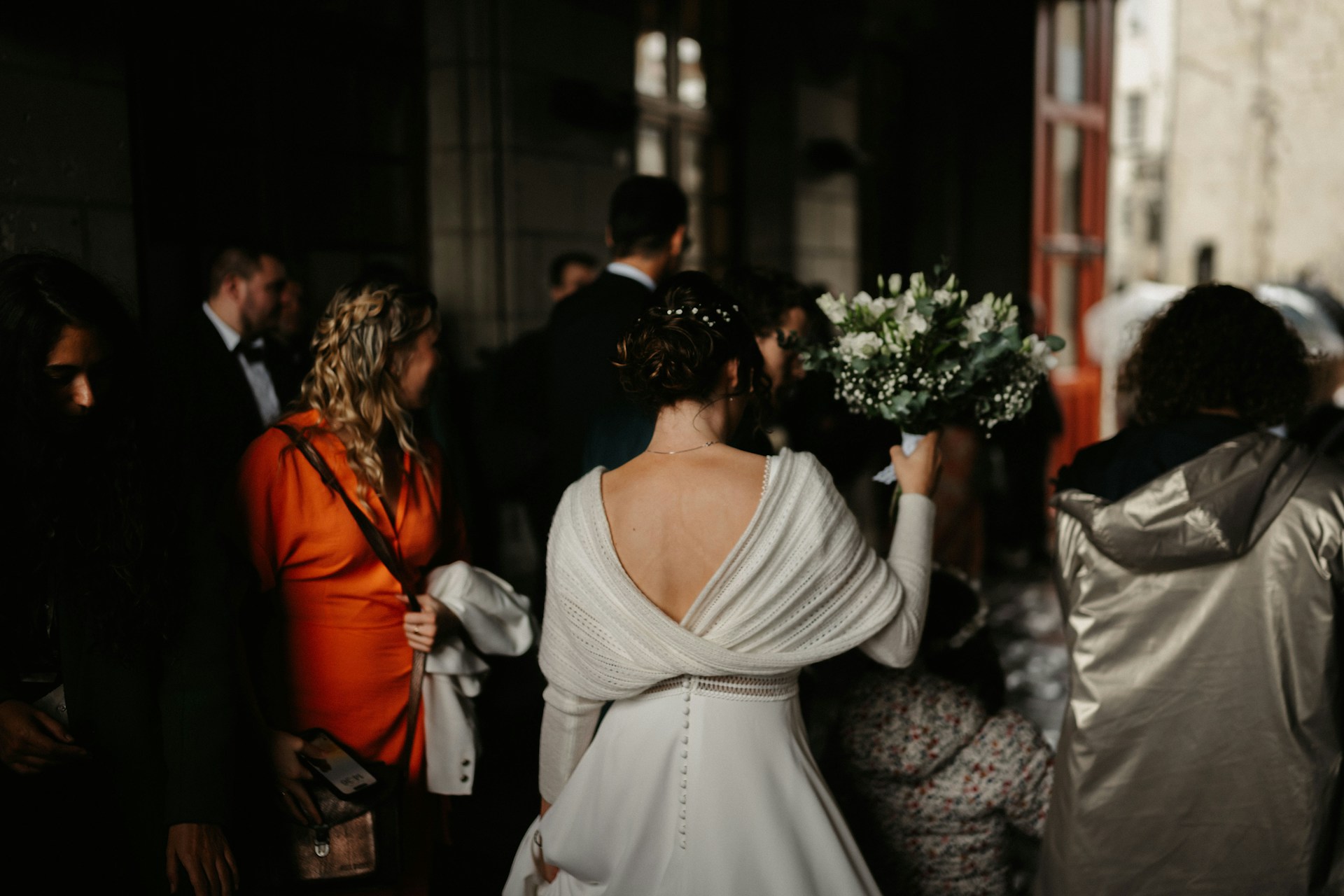 Une mariée de dos tenant son bouquet, entourée d’invités dans une ambiance chaleureuse et tamisée.