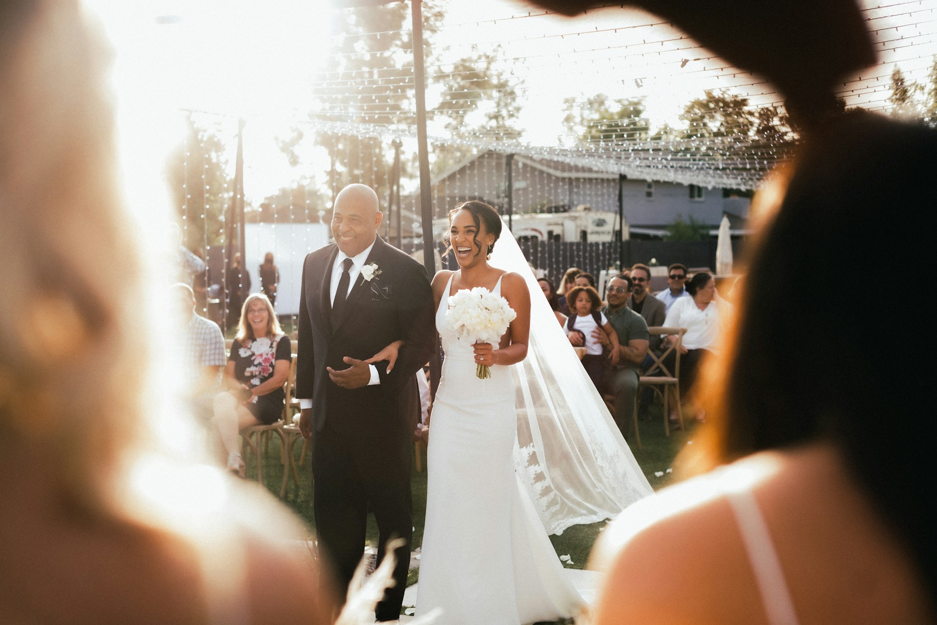 Une mariée radieuse au bras de son père avançant dans l’allée, sous le regard joyeux des invités lors d’un mariage en plein air.